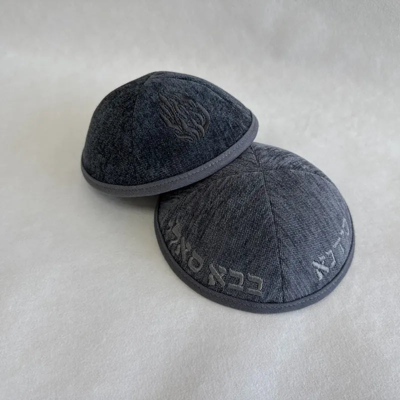 Inscription hébraïque brodée en fil gris sur denim gris, associée à un symbole de flamme sur la face opposée.